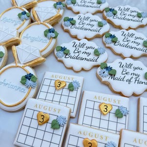 May include: A set of decorated sugar cookies with various designs, including a ring, a calendar with the date August 3rd, and the text "Will you be my bridesmaid?" in different variations. The cookies are decorated with white icing, gold accents, and blue and green floral details.