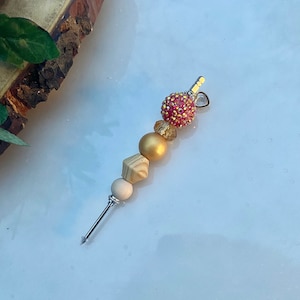 Puede incluir: Un llavero de cuentas de madera y oro con una cuenta rosa brillante y un dije de corazón plateado.