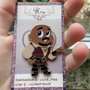 Op de afbeelding: Gouden emaille-pin met een cartoonfiguur met een bruine huid, een bruine baard en een bruin en beige outfit. Het personage draagt ​​een gouden riem met een cirkelvormig ontwerp. De pin is op een witte kaart met de tekst "Lily.B_Pins" en social media-informatie.