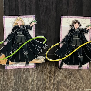 May include: Two enamel pins featuring characters in black cloaks, holding glowing energy whips. One pin has a blonde character with a green whip, the other has a brunette character with a yellow whip. The pins are on decorative cards.