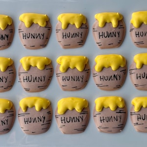 May include: A collection of Winnie the Pooh-themed cookies. Each cookie is shaped like a honey pot, with a brown base and a yellow honey-like topping. The word "HUNNY" is written on each cookie in black.