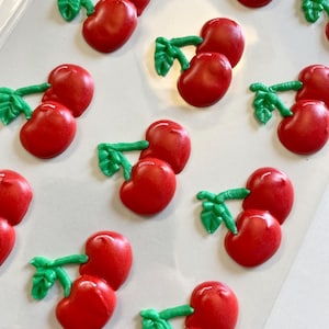 May include: A collection of red cherry-shaped decorations with green stems and leaves. The cherries are glossy and appear to be made of a hard candy or resin material. The image is taken from a top-down perspective.