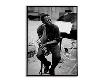May include: Black and white photo of a man sitting on a stool holding a trumpet. He is wearing a dark shirt and jeans. The photo is taken in a studio setting.