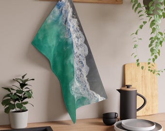 May include: A rectangular kitchen towel featuring a high-resolution aerial print of turquoise ocean waves crashing onto a dark gray sandy beach. The towel is draped over a wooden wall-mounted rack, displaying a vibrant gradient of sea colors.
