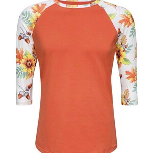May include: A burnt orange baseball tee with a white and floral print on the sleeves. The floral print features sunflowers, leaves, and acorns.