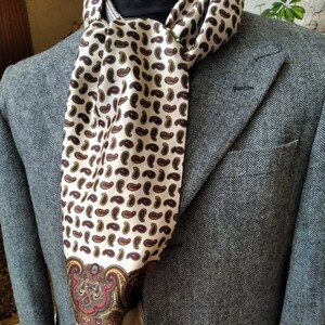May include: A cream-colored silk scarf with a brown paisley pattern is draped over a gray herringbone blazer. The scarf has a decorative border with red and gold accents. The blazer has a notched lapel and a pocket.