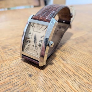 May include: A rectangular Emporio Armani wristwatch with a silver-tone metal case, a light champagne-coloured dial, black Roman numeral hour markers, and a brown textured leather strap with a visible crocodile-style pattern and a metal crown.