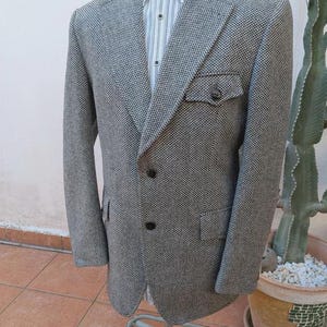 May include: A grey and black speckled tweed blazer featuring a notched lapel, two dark buttons on the front, and a single chest pocket with a buttoned flap. The blazer is worn over a white button-down shirt with thin vertical blue and grey stripes.