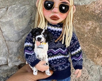 May include: A handmade doll with blonde dreadlocks, sunglasses, and a navy blue sweater with a purple and white pattern. The doll is holding a black and white dog with a tag that says "Sparkle". The doll is wearing ripped blue jeans.