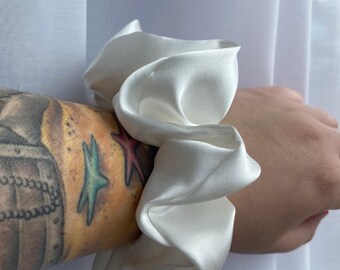 May include: A white satin scrunchie, a hair tie, with a soft, silky texture. The scrunchie is folded and gathered into a large, round shape.