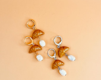 Pode incluir: Um par de brincos de argola de ouro e prata com um charme de croissant e um pingente de pérola. Os brincos estão sobre um fundo laranja claro.