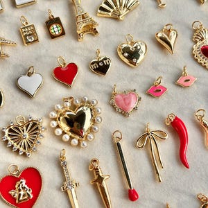 May include: A collection of gold-colored charms with various designs, including hearts, playing card suits, an Eiffel Tower, a fan, and a red chili pepper. Some charms have enamel details in red, white, and pink. The charms are arranged on a light-colored surface.