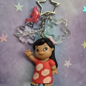 May include: A keychain featuring a figurine of a character in a red dress with white leaf-shaped designs. The keychain includes a star-shaped clasp, a pink fish charm, and clear star and flower-shaped charms. The background is a gradient of purple and blue with star-shaped decorations.