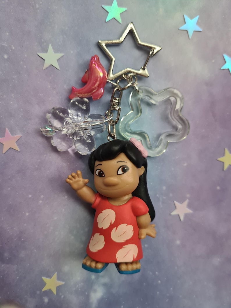 May include: A keychain featuring a figurine of a character in a red dress with white leaf-shaped designs. The keychain includes a star-shaped clasp, a pink fish charm, and clear star and flower-shaped charms. The background is a gradient of purple and blue with star-shaped decorations.
