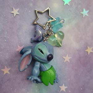 May include: A blue Stitch keychain with a silver star-shaped keyring. The keychain features a Stitch figurine with a green skirt, and charms including a dolphin, a heart, and a four-leaf clover. The background is a purple and blue gradient with star stickers.
