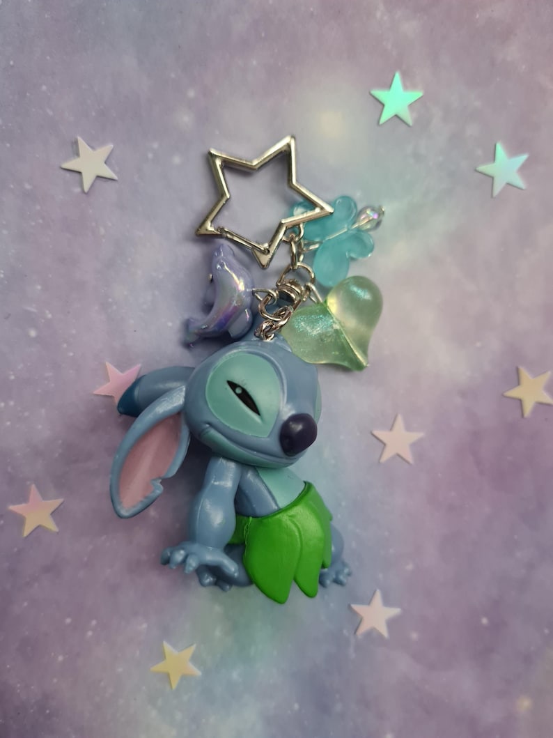 May include: A blue Stitch keychain with a silver star-shaped keyring. The keychain features a Stitch figurine with a green skirt, and charms including a dolphin, a heart, and a four-leaf clover. The background is a purple and blue gradient with star stickers.