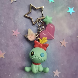 May include: A keychain featuring a light green doll-like figure with a red bow, a star-shaped metal charm, and various charms. The charms include a pink heart, a green star, and a small ice cream cone. The background is a light purple with star-shaped decorations.