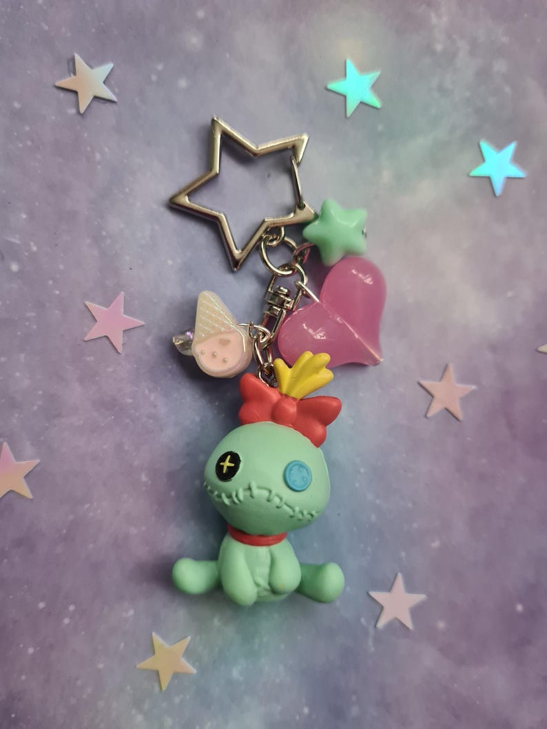 May include: A keychain featuring a light green doll-like figure with a red bow, a star-shaped metal charm, and various charms. The charms include a pink heart, a green star, and a small ice cream cone. The background is a light purple with star-shaped decorations.