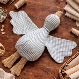 May include: A handmade stuffed goose toy with a blue and white gingham pattern. The wings and legs are a solid tan color. The toy is lying on a wooden surface, surrounded by sewing supplies like scissors, thread, and fabric.