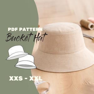 May include: A beige bucket hat with a wide brim sits on a wooden surface. The text "PDF PATTERN Bucket Hat" is displayed above the hat, with "XXS - XXL" below. A line drawing of two bucket hats is on the left.