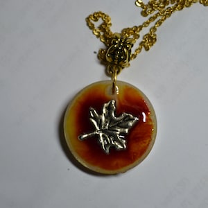 May include: A gold chain necklace with a round pendant featuring a silver maple leaf on a red and yellow background.
