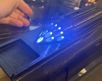 Puede incluir: Un objeto negro impreso en 3D con una luz azul que emana de una abertura circular. El objeto tiene una superficie texturizada y una forma rectangular.