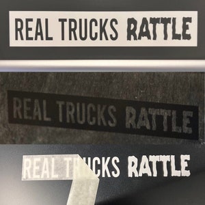 May include: A close-up of a black surface with a white vinyl sticker that reads "REAL TRUCKS RATTLE". The sticker is partially peeled off, revealing the black surface underneath.