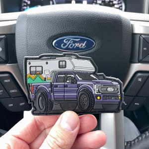 May include: A purple Ford pickup truck with a white camper shell. The truck has black wheels and a black bumper. The camper shell has a window and a door. The truck is on a patch.