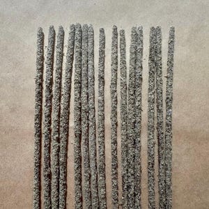 May include: A collection of incense sticks, each with a textured, gray-brown appearance. The sticks are arranged vertically against a light brown background, suggesting a natural or earthy theme. The image is well-lit, highlighting the details of the incense.