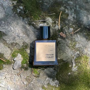 May include: A rectangular perfume bottle with a dark cap and a gold label that reads "OREGON COAST." The bottle is set on a mossy rock surface. The label also includes the text "All natural oils + witch hazel."