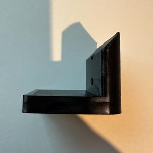 May include: A black, L-shaped bracket with a textured surface. The bracket has a right angle design and appears to be made of plastic or a similar material. The bracket is designed to be mounted on a wall or other surface.