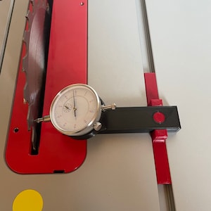 Puede incluir: Un indicador de dial fijado a un soporte negro y rojo se utiliza para medir la alineación de una hoja de sierra de mesa. El indicador de dial lee 0,001 pulgadas.