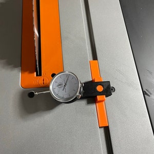 Peut inclure: Un indicateur de cadran fixé à un support noir et orange, mesurant la distance entre une lame de scie et une clôture de scie à table.