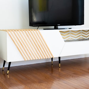 Op de afbeelding: Witte entertainmentcenter met een geometrisch ontwerp aan de voorkant en gouden planken met chevron-patroon. Het entertainmentcenter heeft zwarte poten met gouden tips en staat op een houten vloer.