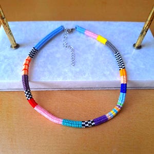 May include: A beaded necklace featuring a vibrant array of colours including light blue, pink, yellow, grey, black, white, orange, purple, red, and teal. The necklace is constructed from small, uniform beads arranged in distinct colour-blocked segments.