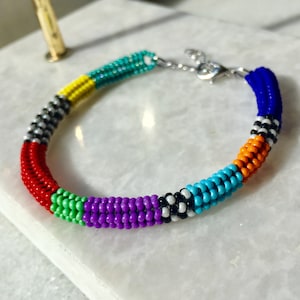 May include: A colorful beaded bracelet with a silver clasp. The bracelet is made of small, round beads in shades of red, orange, yellow, green, blue, purple, black, and white.