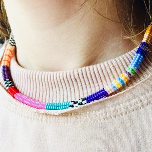 May include: A beaded choker necklace featuring a cylindrical design composed of small, multi-coloured glass beads. The pattern includes segments of solid orange, purple, red, pink, turquoise, blue, yellow, and white, interspersed with black and white accents.