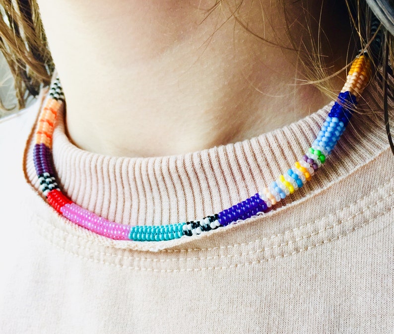 May include: A beaded choker necklace featuring a cylindrical design composed of small, multi-coloured glass beads. The pattern includes segments of solid orange, purple, red, pink, turquoise, blue, yellow, and white, interspersed with black and white accents.