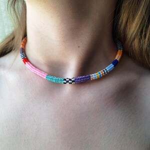 May include: A choker necklace made of small, multi-coloured seed beads arranged in a continuous, cylindrical pattern. The beads feature segments of pink, turquoise, black and white checkered, purple, light blue, dark blue, and orange colours.