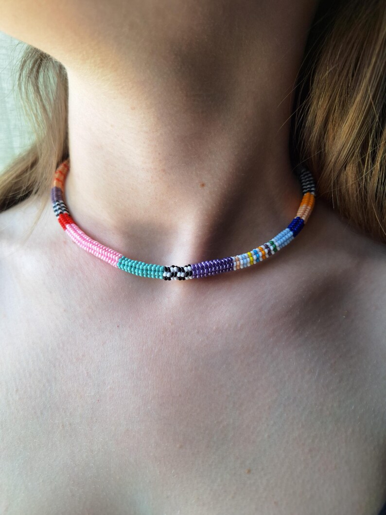 May include: A choker necklace made of small, multi-coloured seed beads arranged in a continuous, cylindrical pattern. The beads feature segments of pink, turquoise, black and white checkered, purple, light blue, dark blue, and orange colours.