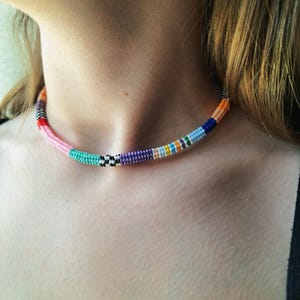 May include: A beaded choker necklace featuring a vibrant array of colours including pink, turquoise, purple, orange, blue, and white. The design incorporates segments of solid colours alongside a small section of black and white chequered beads.