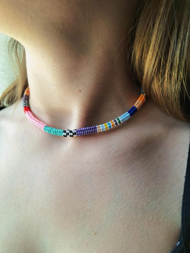 May include: A beaded choker necklace featuring a vibrant array of colours including pink, turquoise, purple, orange, blue, and white. The design incorporates segments of solid colours alongside a small section of black and white chequered beads.