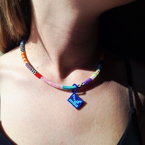 May include: A beaded necklace featuring a multi-coloured cord with segments of orange, pink, turquoise, purple, and black and white chequered patterns. A small, square, blue beaded pendant with a darker blue geometric design hangs from the centre.