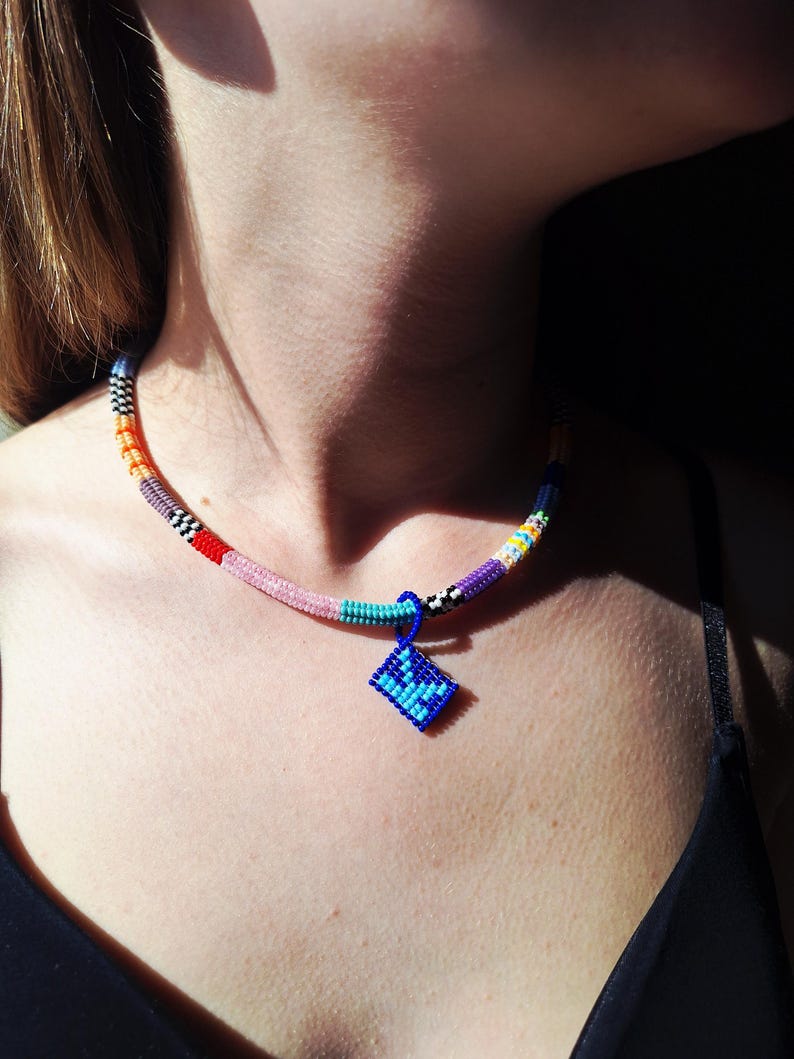 May include: A beaded necklace featuring a multi-coloured cord with segments of orange, pink, turquoise, purple, and black and white chequered patterns. A small, square, blue beaded pendant with a darker blue geometric design hangs from the centre.