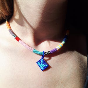 May include: A beaded necklace featuring a multi-coloured cylindrical strand with segments of orange, grey, red, pink, turquoise, black and white, purple, and yellow. A square blue pendant with a darker blue geometric pattern hangs from the centre.