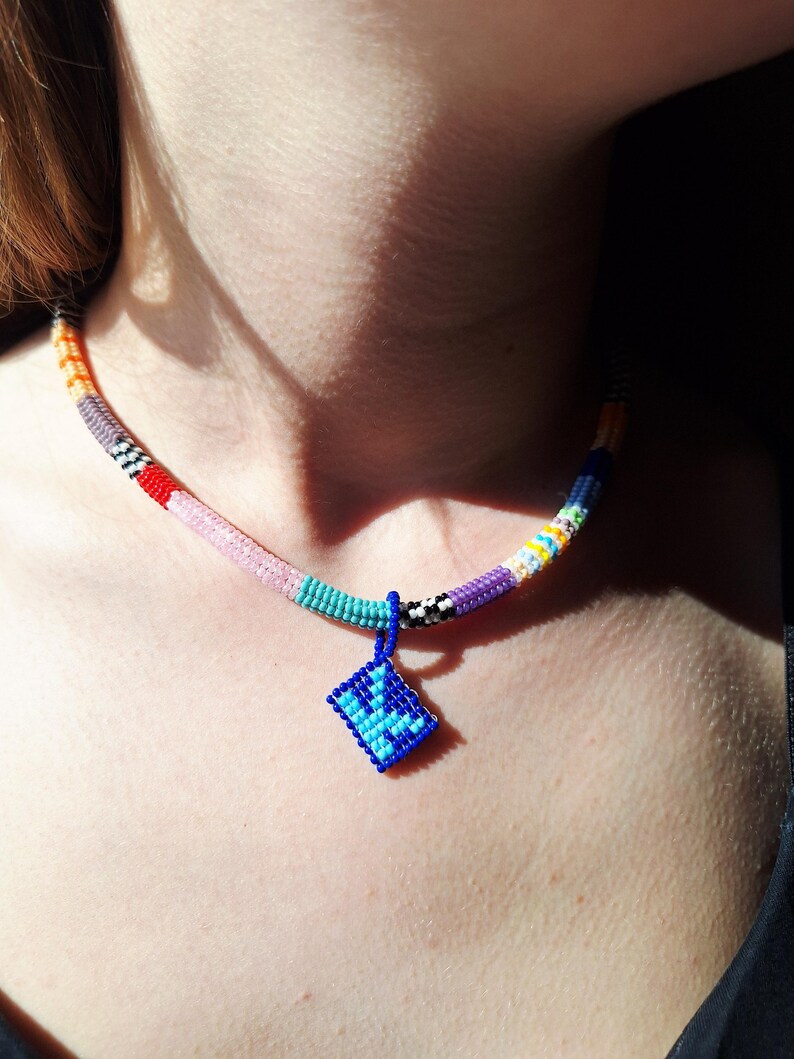 May include: A beaded necklace featuring a multi-coloured cylindrical strand with segments of orange, grey, red, pink, turquoise, black and white, purple, and yellow. A square blue pendant with a darker blue geometric pattern hangs from the centre.