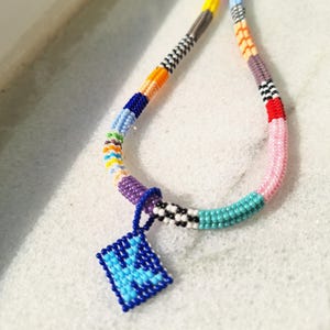 May include: A beaded necklace featuring a multi-coloured pattern of small glass beads in shades of blue, pink, yellow, orange, purple, and white. A square-shaped pendant with a blue and light blue geometric design hangs from the centre.