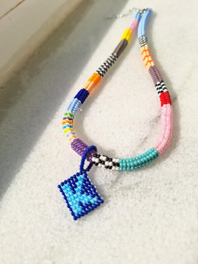 May include: A beaded necklace featuring a multi-coloured pattern of small glass beads in shades of blue, pink, yellow, orange, purple, and white. A square-shaped pendant with a blue and light blue geometric design hangs from the centre.