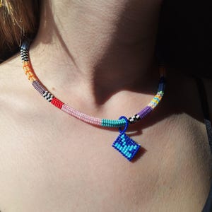 May include: A beaded choker necklace featuring a multi-coloured pattern of orange, pink, teal, purple, and black and white segments. A small, square, blue and light blue beaded pendant hangs from the centre of the necklace on a blue loop.
