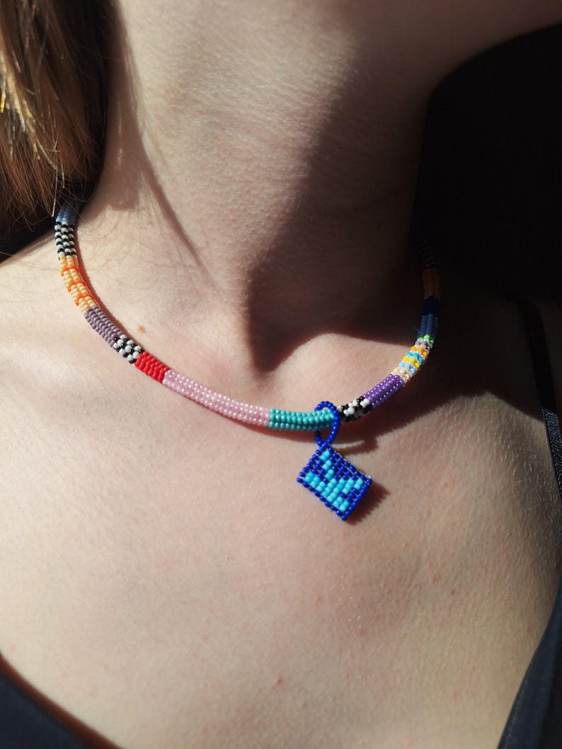 May include: A beaded choker necklace featuring a multi-coloured pattern of orange, pink, teal, purple, and black and white segments. A small, square, blue and light blue beaded pendant hangs from the centre of the necklace on a blue loop.
